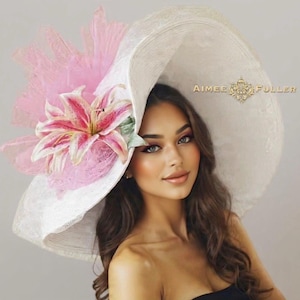 May include: A large, white sun hat with a wide brim, adorned with a pink lily and tulle. The hat features a decorative gold emblem and the text "AIMEE FULLER". This elegant hat is perfect for special occasions.