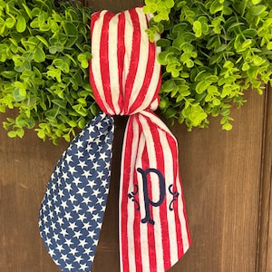 May include: A patriotic wreath decoration featuring a green leafy base and a red, white, and blue ribbon. The ribbon has a striped pattern and a blue section with white stars. A blue letter "P" is embroidered on the ribbon.