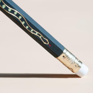 May include: Close-up of a dark teal pencil with a gold snake design and a red tongue. The pencil has a gold ferrule and a white eraser. The snake design features small green dots.