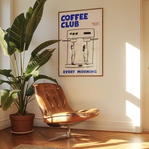 May include: A framed poster with the words "COFFEE CLUB" in blue, above a black and white illustration of a coffee machine and two figures. The words "EVERY MORNING" are below the illustration. A brown leather chair and a potted plant are in the foreground.