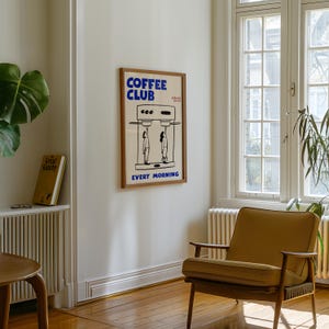 May include: A framed poster with the words "COFFEE CLUB" in blue, above a black and white illustration of a coffee machine and two figures. The text "EVERY MORNING" is below the illustration. The poster is in a room with a chair and a window.