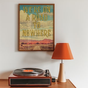 May include: A framed print with a vintage aesthetic featuring a desert landscape with a dirt road leading into the distance. The text "We're on a road to nowhere" is printed in yellow on a blue background.