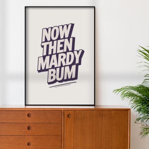 May include: A framed art print with the words "NOW THEN MARDY BUM" in a bold, stylized font. The text is in a dark purple color against a light background. The print is displayed on a wooden cabinet.