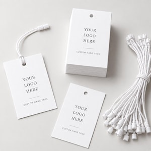 May include: A collection of rectangular white paper hang tags featuring a pre-punched hole at the top. Each tag displays the black printed text YOUR LOGO HERE and CUSTOM HANG TAGS. A bundle of white strings with plastic fasteners is included for attachment.