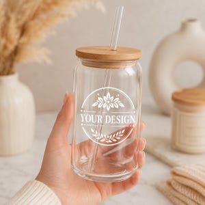 May include: A clear glass tumbler with a bamboo lid and a glass straw. The front of the glass features a white circular graphic with a floral design at the top and bottom, surrounding the centered white text that reads YOUR DESIGN.