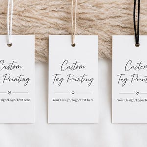 May include: Three rectangular white paper tags with rounded corners, each featuring black text that reads Custom Tag Printing, a small heart icon, and the phrase Your Design/Logo/Text here. Each tag has a different colored string attached.