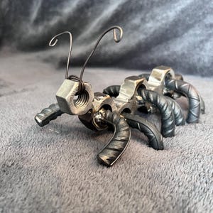 Könnte beinhalten: Eine Metallskulptur einer Ameise aus Muttern, Schrauben und Draht. Die Ameise steht auf einer grauen Oberfläche.