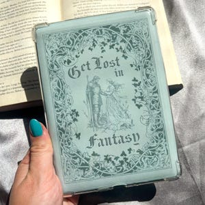 Può includere: Una custodia per tablet azzurra con le parole "Get Lost in Fantasy" in un carattere decorativo. La custodia presenta un'illustrazione di un cavaliere e una donna, circondati da disegni floreali. La custodia è tenuta sopra un libro aperto.