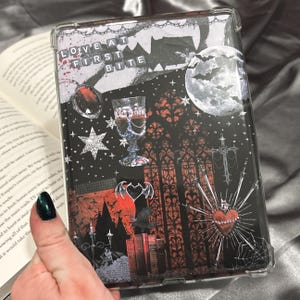 Può includere: Una custodia rettangolare per e-reader con un design a collage e il testo LOVE AT FIRST BITE. La grafica include una luna piena, pipistrelli, un calice, un cuore rosso con punte, una croce, libri ed elementi architettonici gotici in nero e rosso.