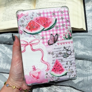 Pode incluir: Uma capa de tablet rosa e branca com um design de fatia de melancia, borboleta e laço. A capa apresenta um padrão floral e xadrez, com corações e estrelas. A capa é segurada em uma mão.