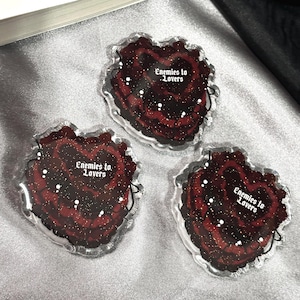May include: Three heart-shaped acrylic pins featuring a dark red and black layered design with gold glitter accents. Each pin displays the white text Enemies to Lovers in a gothic font style, set against a deep red heart with black edges.