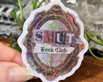 May include: A sticker with a decorative frame design and the words "SMUT Book Club" in a mix of fonts. The sticker has a white border and is covered in iridescent glitter.