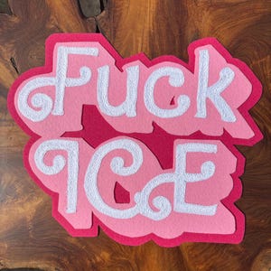 May include: A layered felt wall hanging featuring the text "FUCK ICE" in white, textured, cursive-style lettering. The letters are set against a light pink background layer, which is framed by a larger, dark magenta felt backing piece.