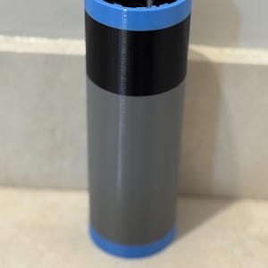 May include: A cylindrical object with a blue top and bottom, a black band in the middle, and a gray body. The top has a ridged design. The object is approximately 6 inches tall.