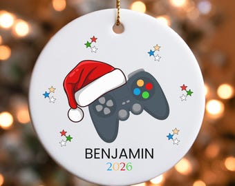 May include: A round white ceramic ornament featuring a dark gray video game controller wearing a red and white Santa hat. The design includes small colorful stars and the text BENJAMIN 2026 printed in black and multicolored numbers below the controller.
