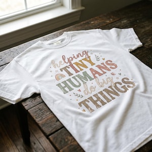 May include: White t-shirt featuring the phrase "helping TINY HUMANS do big THINGS" in a retro font with a rainbow and stars. The design uses pastel colours. The t-shirt is displayed on a wooden table.