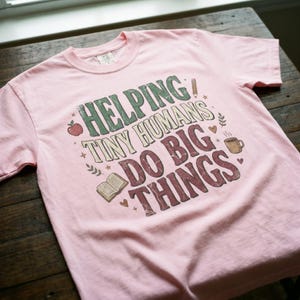 Puede incluir: Una camiseta rosa claro con el texto "HELPING TINY HUMANS DO BIG THINGS" en verde, crema y granate. El diseño incluye una manzana, un lápiz, un libro, una taza de café y otros pequeños gráficos. La camiseta es de material suave.