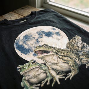 Può includere: T-shirt nero con una dettagliata illustrazione di tre rane davanti a una luna piena. Le rane sono in tonalità di verde e marrone, con la luna in blu e bianco. La maglietta è in materiale morbido.