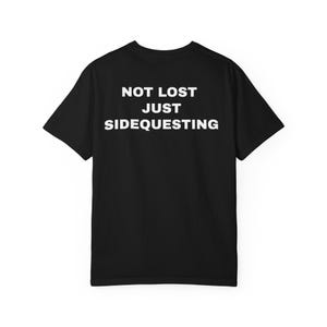 Könnte beinhalten: Schwarzes T-Shirt mit dem weißen Text "NOT LOST JUST SIDEQUESTING" auf dem Rücken. Das Shirt hat kurze Ärmel und einen Rundhalsausschnitt. Der Text ist zentriert und in einer fetten, serifenlosen Schrift.