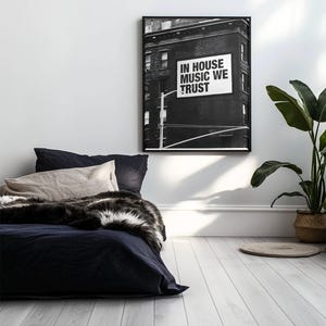 May include: Black and white framed art print featuring a building with a sign that reads "IN HOUSE MUSIC WE TRUST." The print hangs above a bed with navy blue bedding, a beige pillow, and a faux fur throw. A potted plant sits in the corner.