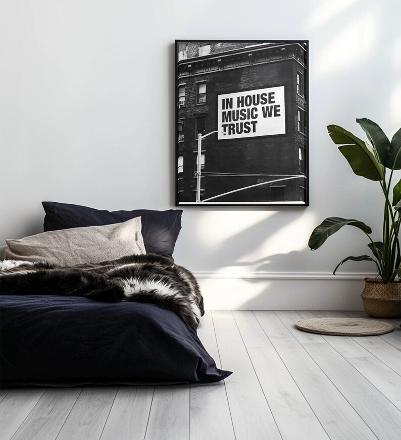 May include: Black and white framed art print featuring a building with a sign that reads "IN HOUSE MUSIC WE TRUST." The print hangs above a bed with navy blue bedding, a beige pillow, and a faux fur throw. A potted plant sits in the corner.