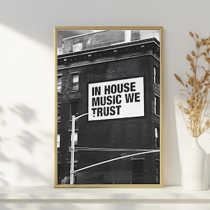May include: Black and white framed print featuring a building with a sign that reads "IN HOUSE MUSIC WE TRUST." The image is displayed on a white shelf with a white vase and dried plants. The frame is gold.