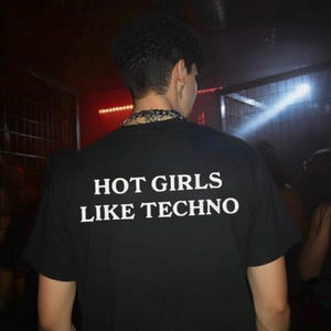 May include: Black t-shirt with the white text "HOT GIRLS LIKE TECHNO". The shirt is worn by a person with dark curly hair and a silver chain necklace. The background is dark with red and white lights.