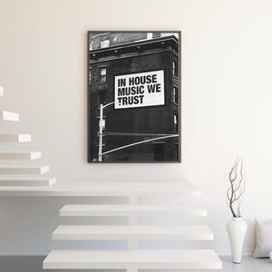 May include: Black and white framed print featuring a building with a sign that reads "IN HOUSE MUSIC WE TRUST." The artwork is displayed on a white wall above a white staircase. A white vase with branches and a pillow are on the floor.