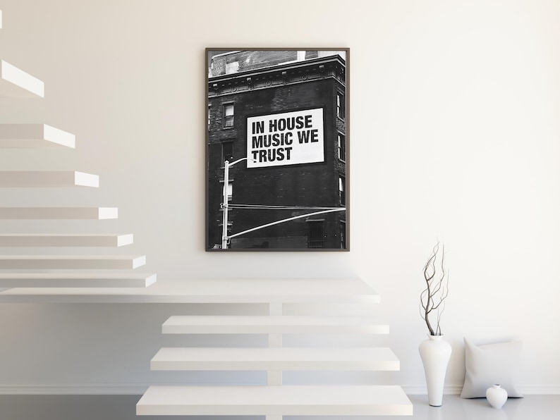 May include: Black and white framed print featuring a building with a sign that reads "IN HOUSE MUSIC WE TRUST." The artwork is displayed on a white wall above a white staircase. A white vase with branches and a pillow are on the floor.