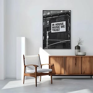 May include: A black and white framed print with the text "IN HOUSE MUSIC WE TRUST" hangs above a wooden cabinet. A mid-century modern chair with a light-colored woven seat and wooden frame sits in front of a white wall.