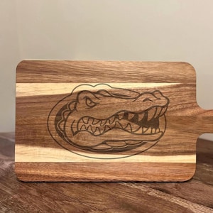 Puede incluir: Tabla de cortar de madera con asa, con un diseño de caimán grabado con láser. La tabla está hecha de tiras de madera claras y oscuras, creando un patrón a rayas. El caimán está detallado con dientes afilados y una expresión amenazante.