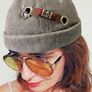 May include: A gray denim cap with a brown leather band adorned with red beads and silver accents. The cap features metal eyelets and a small silver charm. The image also shows aviator sunglasses with orange lenses.