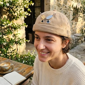 May include: A beige brimless cap featuring a decorative metal butterfly ornament attached to the front. The butterfly is constructed from four distinct textured metal discs in gold and silver tones, connected by small silver rings and thin metal wires.