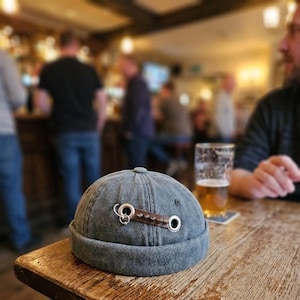 May include: A grey denim docker cap featuring a brown leather strap detail across the front. The strap is secured with two large silver-toned metal grommets and includes a small silver ring attachment and a series of small metal studs.