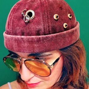 May include: A burgundy velvet beanie hat with metal grommets and decorative studs. The hat features a rolled brim and is accessorized with a silver hoop earring. The person is wearing aviator sunglasses with orange lenses.