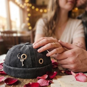 May include: A charcoal gray brimless cap featuring a silver-toned metal key charm engraved with the word Love, attached to a circular metal ring. The cap includes additional gold-toned metal eyelets along the side of the fabric crown.