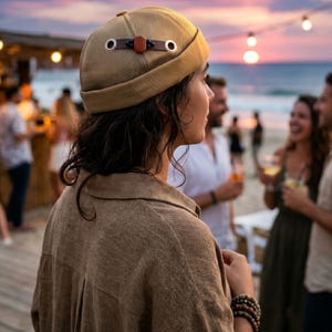 May include: A tan, brimless docker cap featuring a mesh crown, a solid fabric band, and a decorative brown leather strap across the side with two silver-toned metal grommets and a central dark brown accent piece.