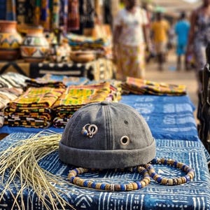 May include: A charcoal gray brimless cap featuring a silver metal ring and a small blue bead attached to the side, along with a silver metal eyelet. A strand of blue, tan, and brown wooden beads is positioned in front of the cap.