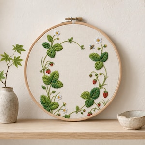 May include: A circular wooden embroidery hoop containing off-white fabric with a hand-stitched design of green strawberry leaves, small white flowers, red strawberries, and a tiny bee, arranged in a wreath pattern around the center.