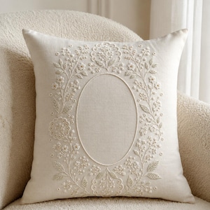 May include: A square off-white throw cushion featuring intricate cream-coloured embroidery. The design showcases a central oval frame surrounded by detailed floral and botanical patterns, including small leaves, buds, and scalloped circular motifs.