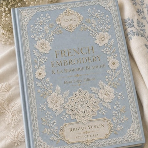 May include: A light blue hardcover book titled French Embroidery & La Broderie Blanche Book 2 by Ridvan Yusein Stitchesly. The cover features intricate white lace-like floral patterns, ornate borders, and gold lettering on a matte blue background.