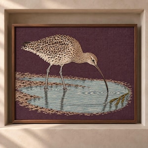 May include: A framed embroidery art piece featuring a brown and tan speckled curlew bird standing in a shallow pool of blue water. The bird has a long, curved beak touching the water surface, creating ripples against a deep plum-coloured background.