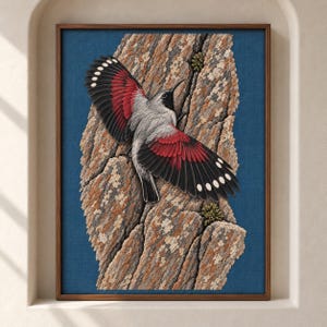 Op de afbeelding: Een ingelijst kunstwerk aan de muur met een gedetailleerd borduurwerk van een vogel met zwart-wit-rode vleugels, zittend op een gestructureerde, lichtbruine boomstam. De achtergrond is diepblauw en het geheel zit in een houten lijst.