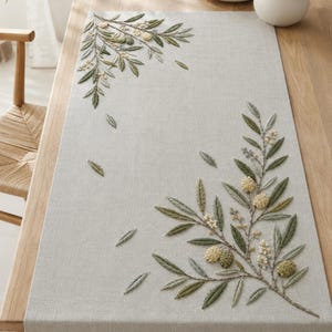May include: A light grey rectangular table runner featuring detailed embroidery of olive branches with green leaves, small white flowers, and textured olive fruits. The embroidery is positioned in the top-left and bottom-right corners of the fabric.