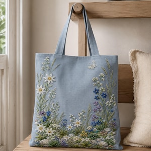 May include: A light blue tote bag featuring intricate embroidery of white edelweiss flowers, blue wildflowers, green foliage, and a small white butterfly. The floral design is arranged along the bottom and sides of the fabric bag.