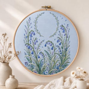 Op de afbeelding: Een ronde houten borduurring met lichtblauwe stof en handgeborduurde klokjesbloemen in blauwe, paarse en groene tinten. Het borduurwerk toont hoge stengels, fijne bladeren en een gebogen schikking van kleine bloemknoppen.