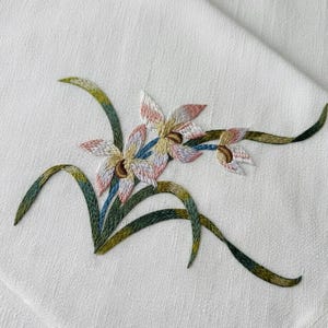 May include: Embroidered floral design on white fabric. The flowers have pink, beige, and white petals with brown centers. Green and blue stems and leaves curve across the fabric, creating a delicate, handcrafted look.