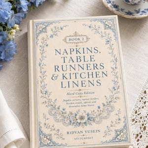 May include: A hardcover book titled Book 2 Napkins, Table Runners & Kitchen Linens by Ridvan Yusein. The cover is cream-colored with blue floral borders and text. It features subtext about napkin corners, runner borders, and kitchen linens.