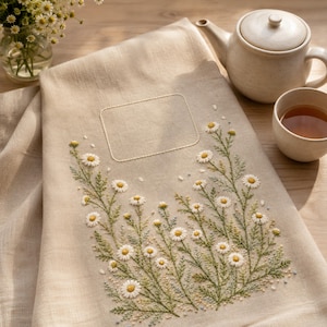 May include: A beige linen tea towel featuring an embroidered design of white and yellow daisy flowers with green stems and leaves. The towel includes a rectangular stitched frame above the floral arrangement, providing a space for text.