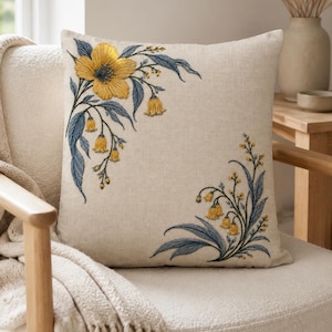 Puede incluir: Un cojín cuadrado con una base de tela beige claro que presenta un bordado intrincado en las esquinas superior izquierda e inferior derecha. El diseño muestra flores amarillas y hojas azules con tallos, creando un patrón floral.